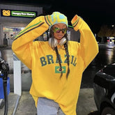 Person wearing a yellow 'Brazil' sweatshirt with braided hair, standing at a gas station.